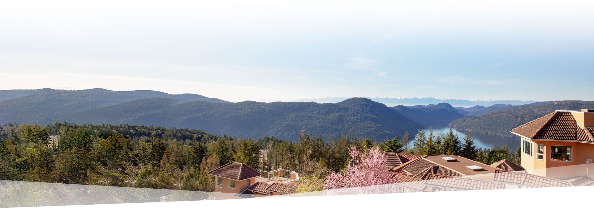 A scenic view of mountainous terrain under a clear blue sky. In the foreground are buildings with red-tiled roofs, surrounded by evergreen trees and blooming pink flowers. A distant lake reflects the sky.