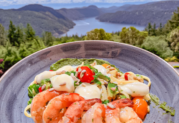 A plate of shrimp pasta with cherry tomatoes and mozzarella, garnished with pesto, is set against a stunning outdoor backdrop of a forested valley and distant lake under a clear blue sky.