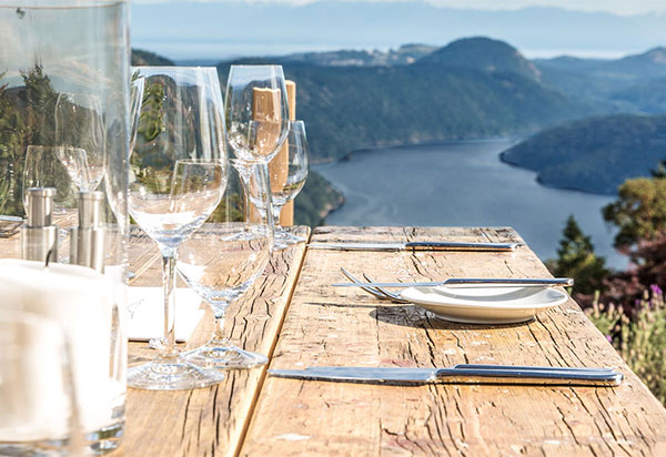 An outdoor table setup with wine glasses and water glasses with the Saanich inlet in the background.